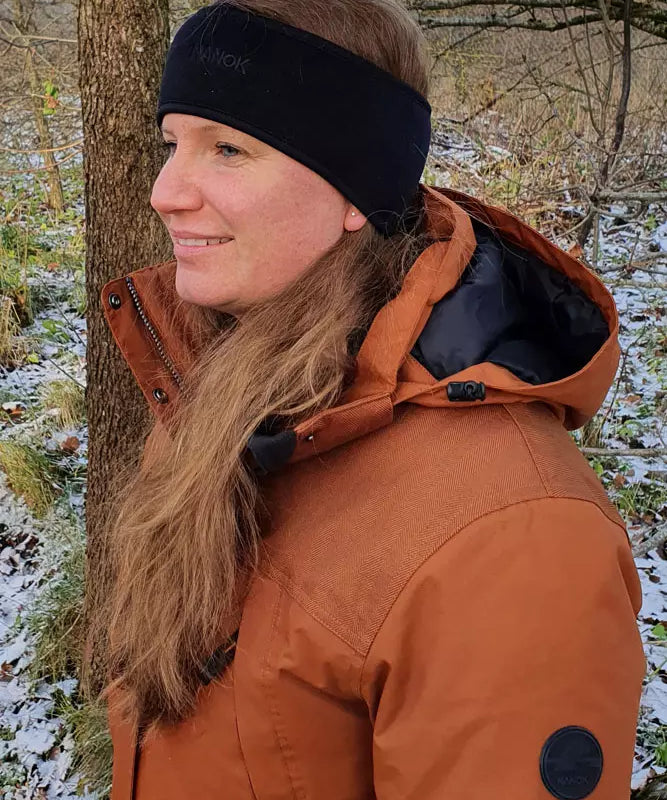 En kvinde står i en let snedækket skov og smiler roligt, mens hun ser ud mod horisonten. Hun har langt, lyst hår samlet bag et sort pandebånd og bærer en varm, orange vinterjakke med hætte. Det naturlige miljø med træstammer, visne blade og strejf af sne skaber en stemningsfuld og fredfyldt vinteragtig atmosfære.