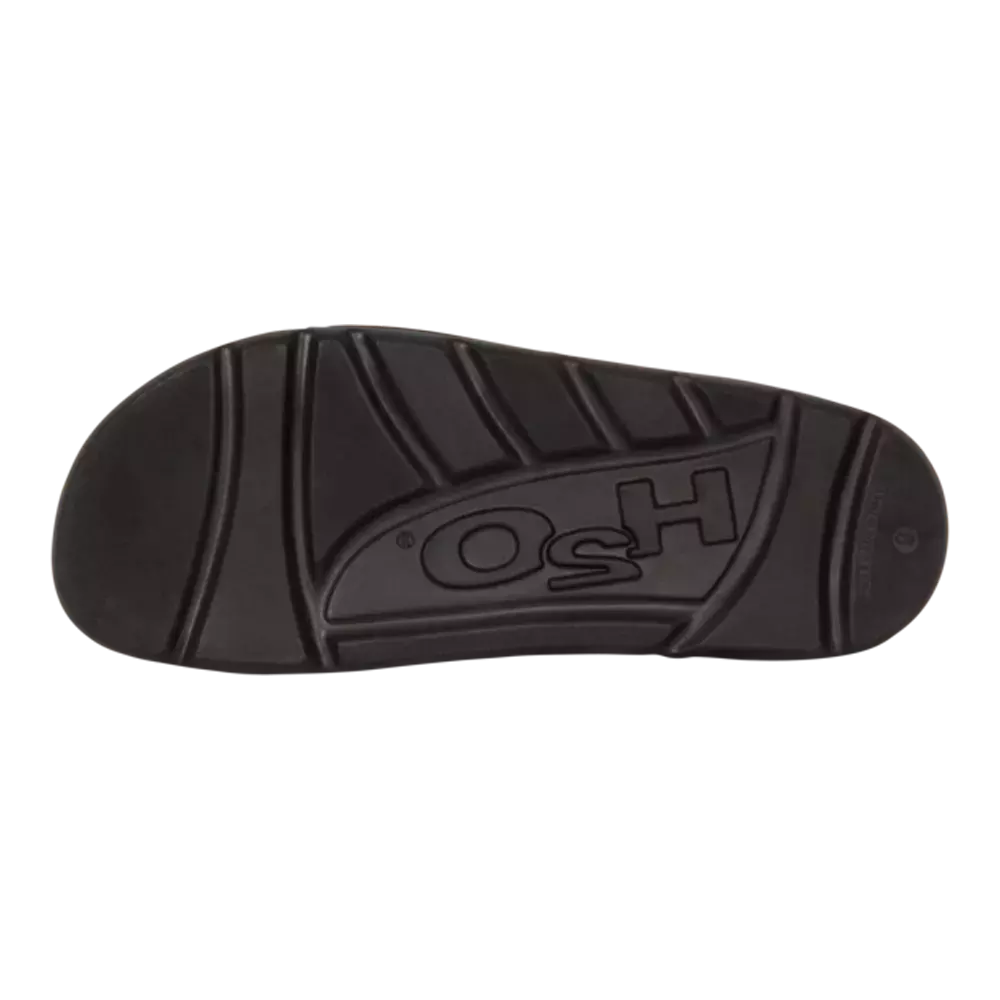 Billedet viser en sort gummisål set nedefra med markerede slidbaner og dybe riller for godt greb. Sålens asymmetriske mønster kombinerer brede baner og skrå spor, hvilket giver øget stabilitet og afvanding. Materialet virker robust og støddæmpende, egnet til hverdagssko eller arbejdstøj. Et indgraveret logo ses i midten, hvilket antyder en færdigvare fra en etableret producent.