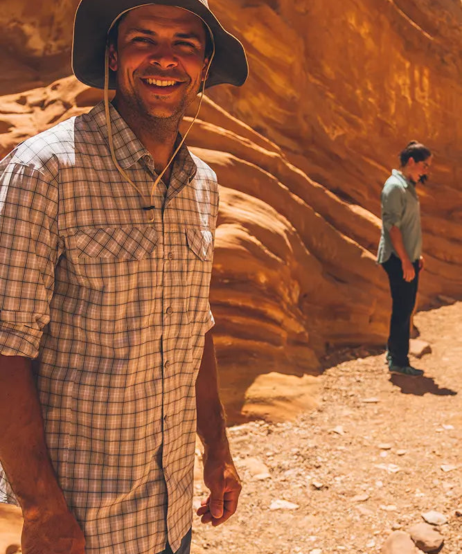 En mand i forgrunden smiler i en ternet skjorte og bredskygget hat, badet i varmt sollys, mens han står i en snæver, orange sandstenskløft. Den rødlige klippevæg danner bølgende mønstre bag ham. En anden person står i baggrunden og kigger væk langs stien, hvilket giver billedet en følelse af rolig opdagelse og ørkenens stille, barske skønhed.