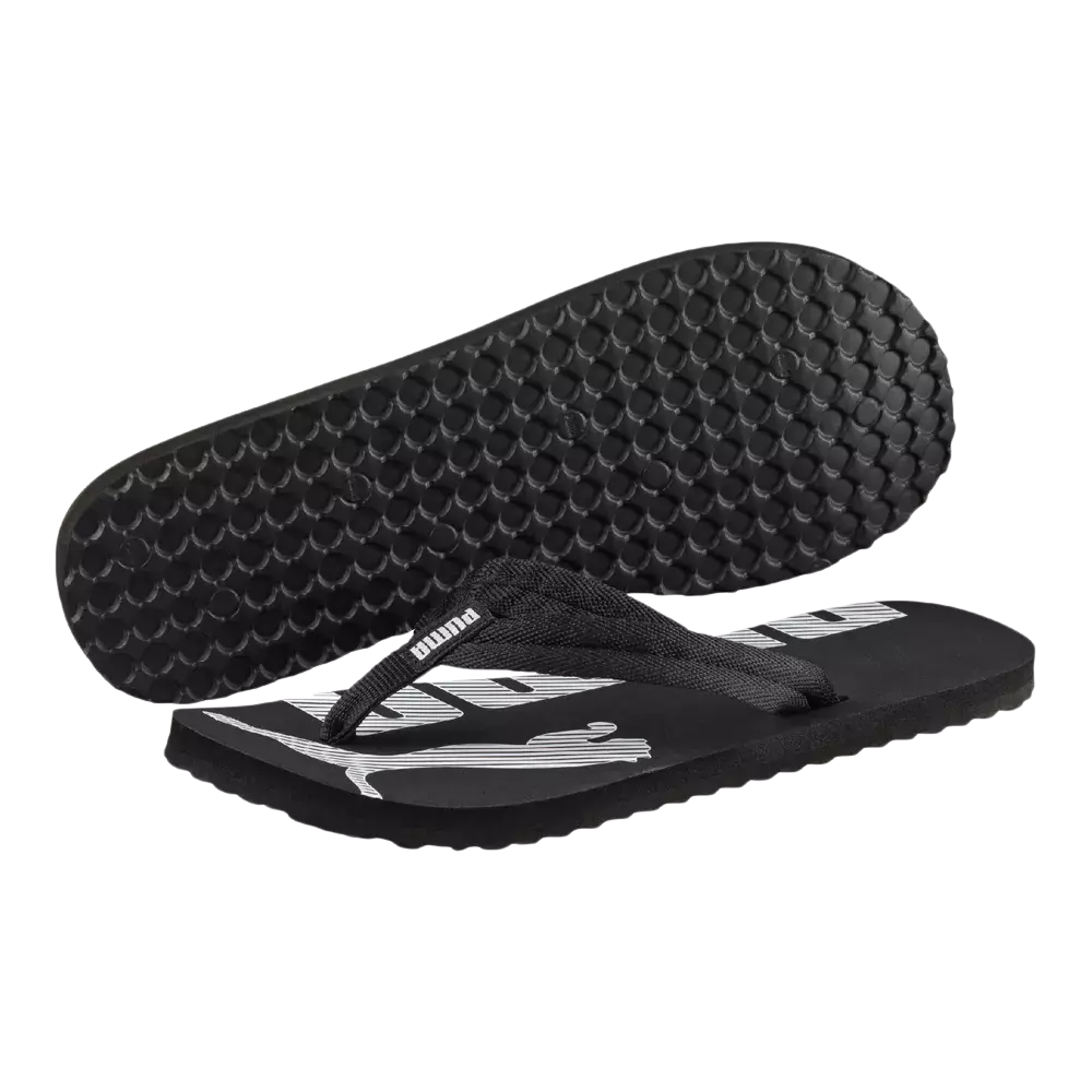 På billedet ses et par sorte flipflops med brede tekstilremme og en flad, let sål. Overfladen prydes af et stort, hvidt Puma-logo, mens undersiden har et mønster af små halvkloder for bedre greb. Sandalerne virker simple og funktionelle, velegnede til strand, bad eller afslappet fritid. Materialet ser ud til at være skumgummi med stofstropper, der giver komfort og holdbarhed.
