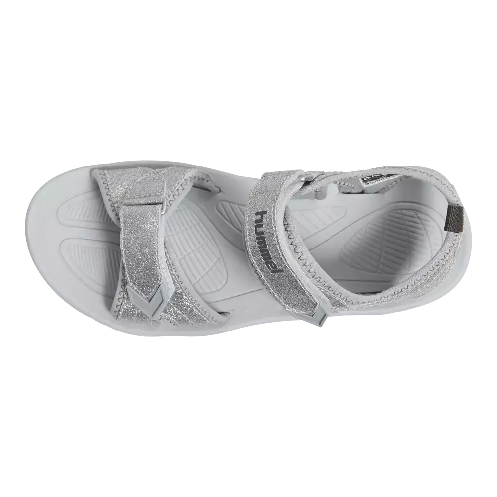 Et enkelt sølvfarvet sportssandal set oppefra med glitrende remme og to justerbare velcrolukninger. Den bløde, formstøbte indersål har mønstrede fodaftryk for ekstra greb, mens den let profilerede ydersål sikrer stabilitet. Sandalen fremstår både praktisk og pyntelig, velegnet til hverdagsbrug og leg, og kombinerer komfort med et moderne, skinnende finish. Brandets navn ses diskret på remmen.