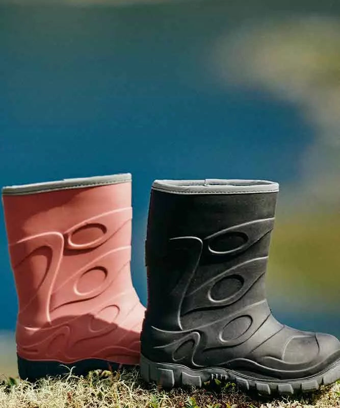 To robuste gummistøvler står side om side på en mosdækket klippe ved vandkanten. Den ene er lyserød med bløde, indstøbte mønstre, den anden er mørkegrå med grov sål og markante strukturer. Baggrunden er sløret blå, hvilket antyder sø eller hav og skaber kontrast til støvlernes farver. Billedet formidler enkelhed, funktionalitet og et lille strejf af udendørs eventyr.