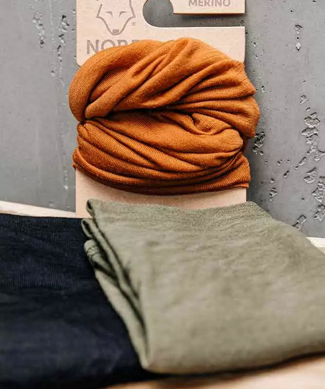 halstørklæde er monteret på en papplade med logo og teksten "100% MERINO". Foran ligger to sammenfoldede stykker stof i olivengrøn og sort på en lys træflade, mod en ru, grå betonbaggrund. Stemningen er nordisk, naturlig og funktionel, velegnet til kølige dage.