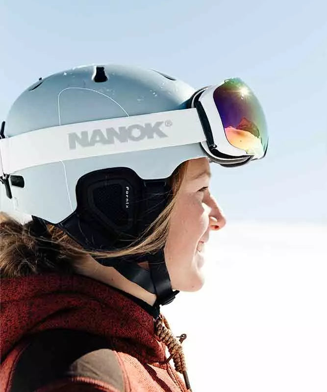 En ung person i profil iført lyseblåt skihjelm med "NANOK"-stroppen og spejlreflekterende skibriller. Personen smiler mod det klare, sneklædte landskab, iført en rødbrun vinterjakke med snøre ved kraven. Hjelmens polstring og hakestrop sidder korrekt, og solen reflekteres i glasset på brillen. Billedet formidler en følelse af frisk luft, glæde og forberedelse til en dag på pisterne.