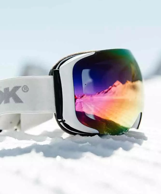 Et par skibriller ligger i sneen, deres reflekterende linse viser et farverigt panorama af bjerge og himmel i nuancer af pink, orange og lilla. Den hvide ramme og stroppen står i kontrast til den spejlblanke overflade, mens blødt lys og skygger falder over sneens tekstur. Billedet formidler en stille vinterstemning og længsel efter friske spor på en solrig fjeldsdag.