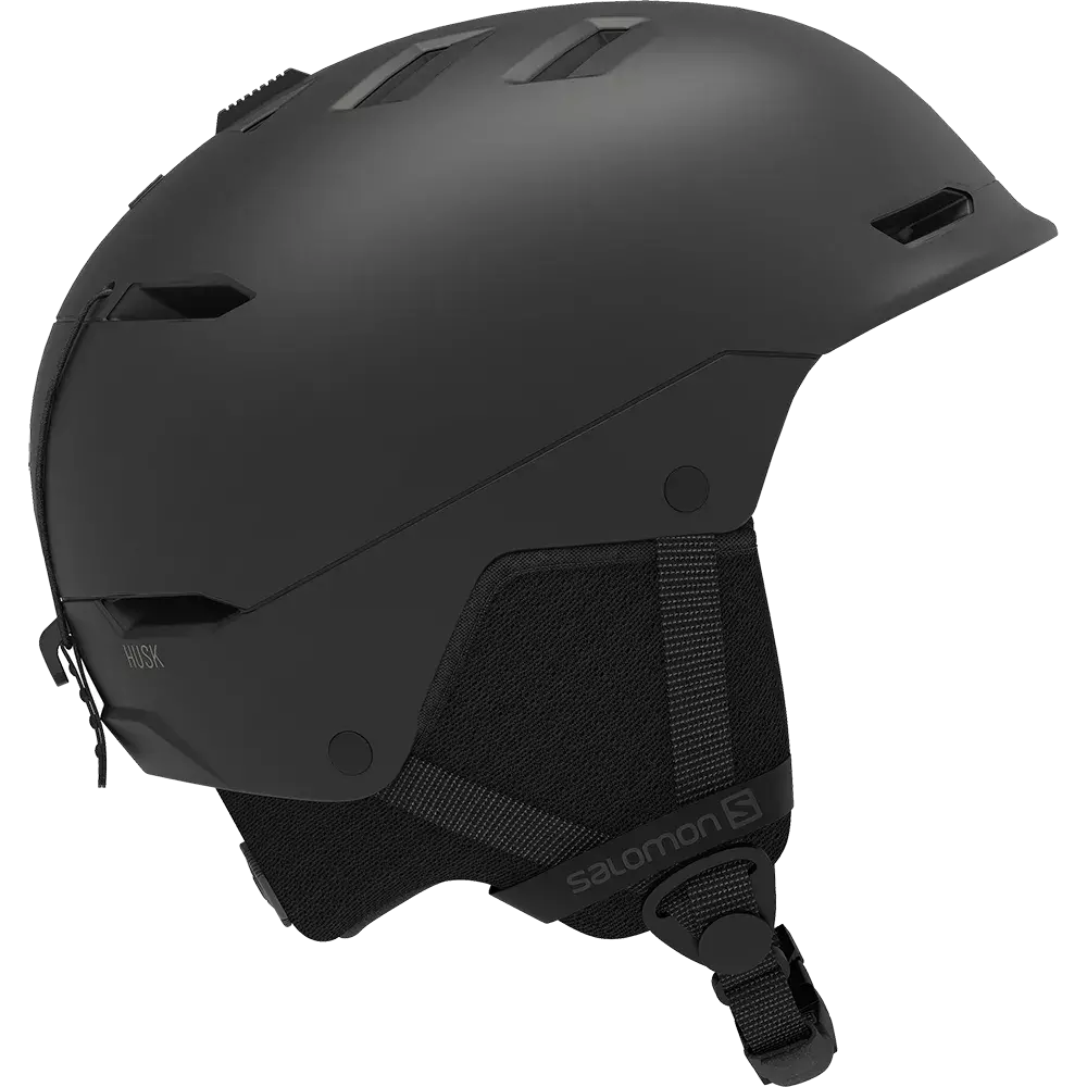 Mat sort ski eller snowboardhjelm med mat overflade og en strømlinet, moderne form. Hjelmen har flere ventilationsåbninger i toppen og på siderne, polstrede øreklapper i tekstil og en justerbar hagerem med klikspænde. Skallen virker robust og støddæmpende, mens samlinger og monteringer er diskrete. Logoet fra Salomon ses på remmen, og designet kombinerer funktionalitet med minimalistisk æstetik.