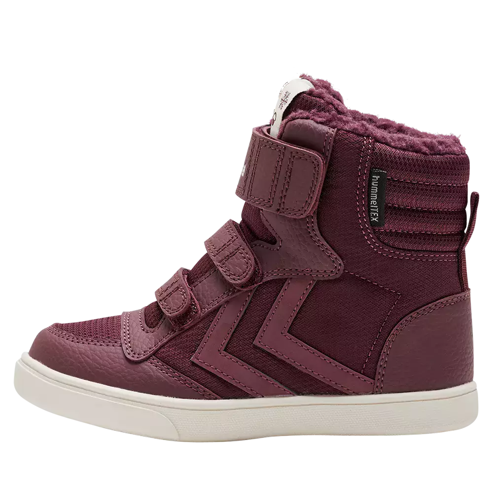 hindbærrødt højtop-sneakersstøvle til børn med blødt, pelsforet skaft. Overdelen kombinerer tekstureret læder og stofpaneler med dekorative syninger og et karakteristisk zigzag-motiv langs siden. Skoen lukkes med tre praktiske velcroremme, og en mærkeetiket "hummelTEX" sidder ved anklen. Den lyse, kraftige gummisål giver god støtte og kontrast, hvilket gør støvlen både varm, komfortabel og velegnet til hverdag.