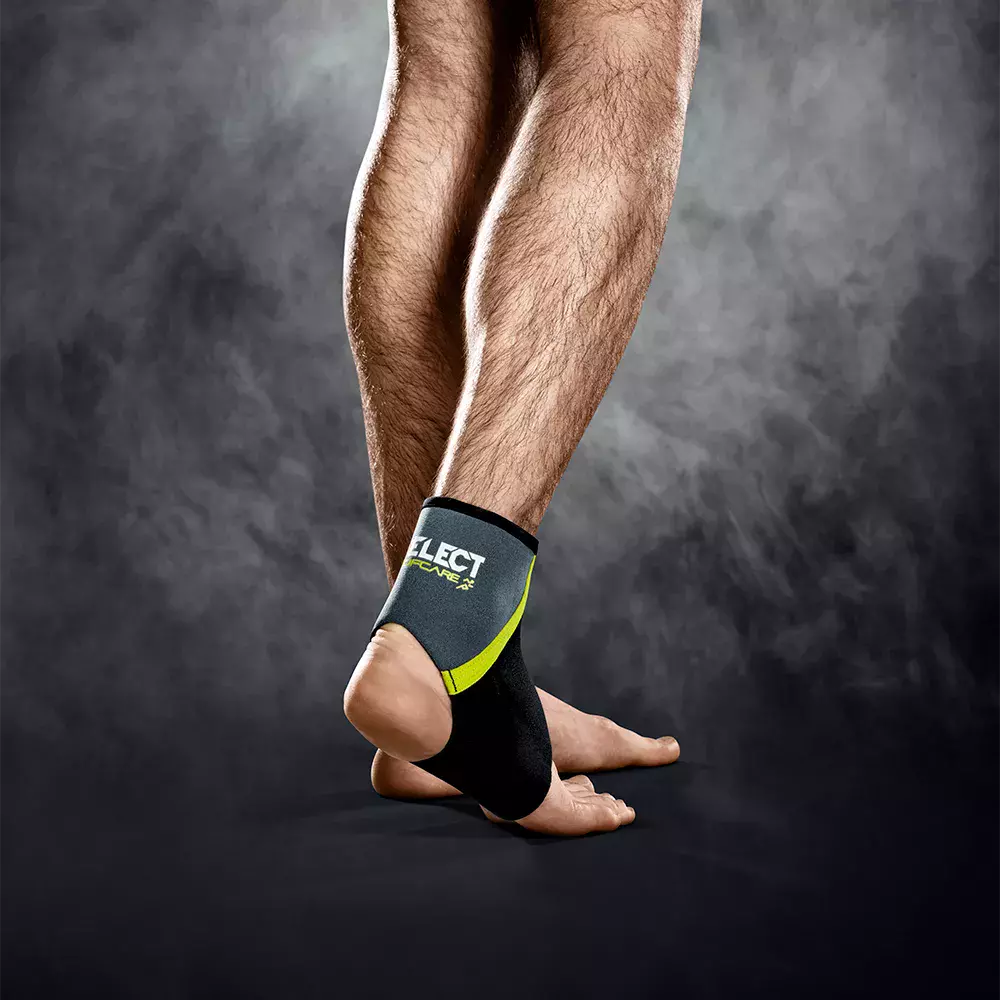 Et nærbillede viser en persons underben og fødder i en atletisk positur, stående på tæerne. Anklen er støttet af en sort-grå ankelbandage med grønne detaljer og teksten "SELECT", designet til stabilitet og beskyttelse. Baggrunden er mørk og let røgfyldt, hvilket fremhæver muskulaturen og produktets funktion som støtte under træning eller genoptræning.