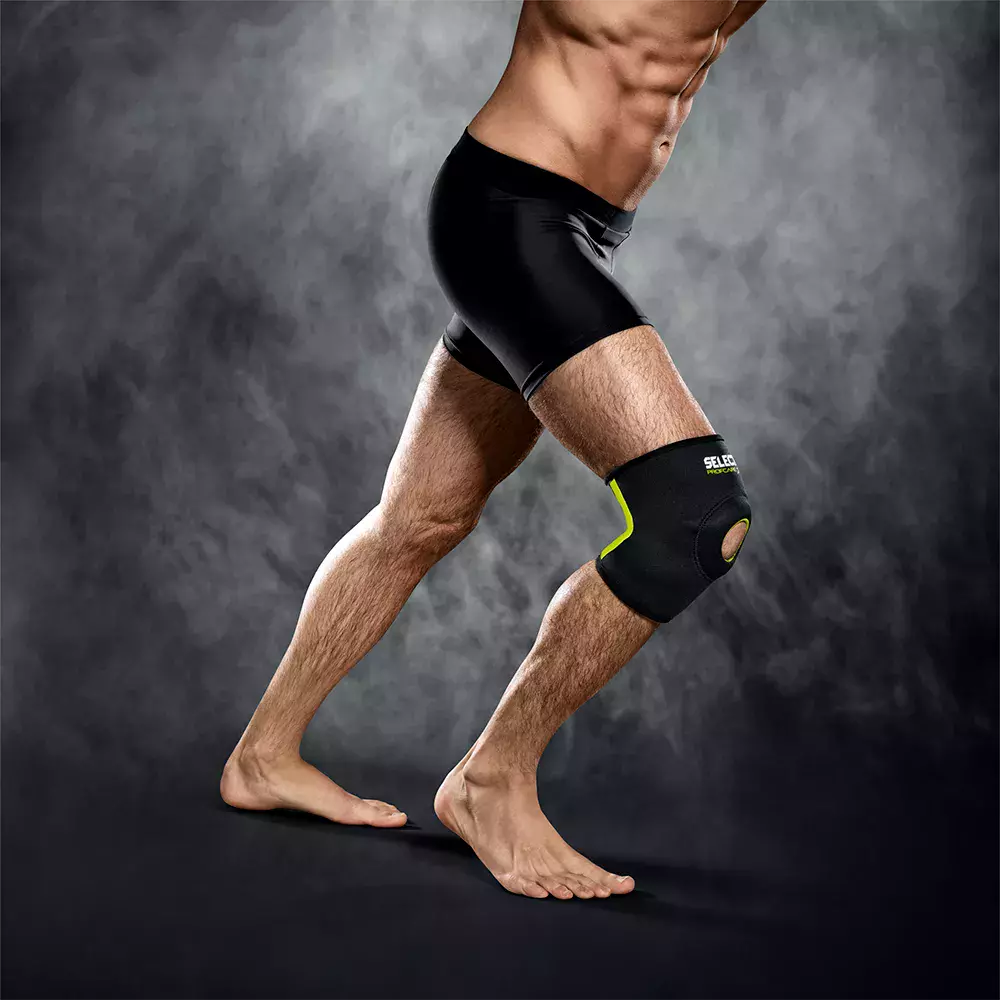 Et dramatisk studie af en atletisk person i knælængde: stærke, veldefinerede ben og en markeret mave i sorte kompressionsshorts. Personen står i et skridt, barfodet, med fokus på højre knæ, der er beskyttet af en sort knæbandage med neongrøn kant og åbning ved knæskallen. Mørk, røgfyldt baggrund og kontrastfuld belysning fremhæver muskulatur og bevægelsesenergi.