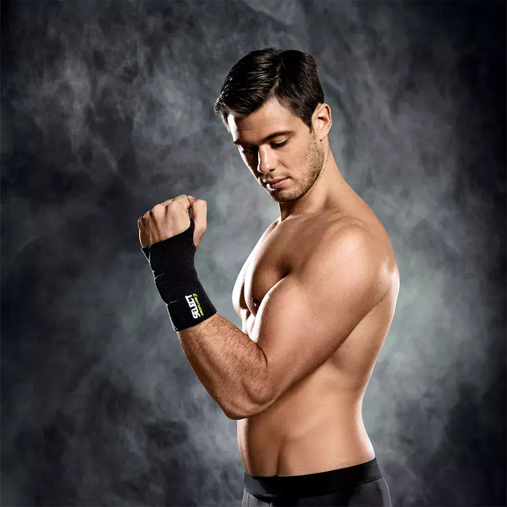 En atletisk, ung mand står uden trøje foran en mørk, røgfyldt baggrund. Han har kort, mørkt hår og viser en veltrænet overkrop, mens han bøjer armen og inspicerer sit håndled. På håndleddet ses en sort støttebind med logo. Billedet har et dramatisk, koncentreret udtryk og signalerer træning, styrke og fokus, som om han forbereder sig til en øvelse eller kamp.