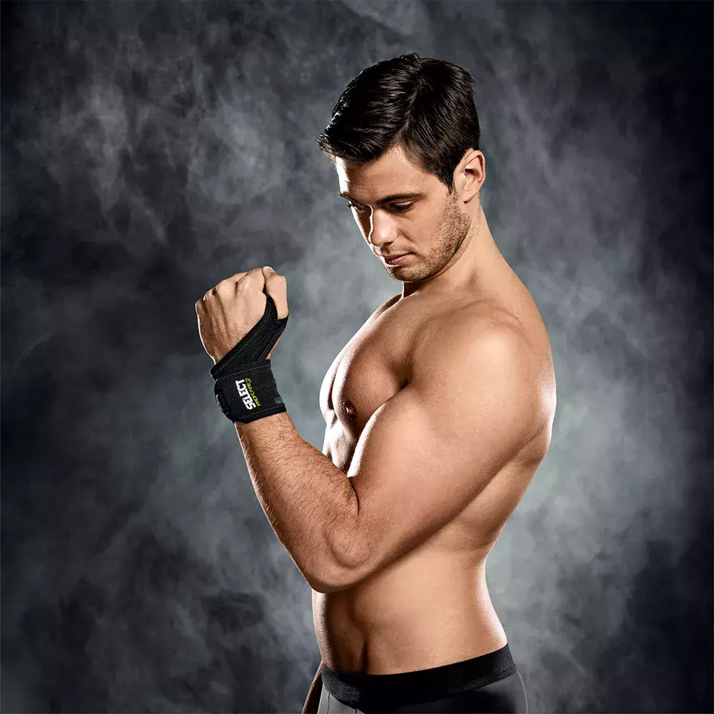 En muskuløs, skjorteløs mand står i profil mod en mørk, røgfyldt baggrund og kigger koncentreret ned på sin håndledsbandage. Hans veltrænede overkrop og markerede arm fremhæver en trænings- og rehabiliteringsstemning. Det enkle sportslige outfit og intensiteten i blikket signalerer fokus, styrke og dedikation til fysisk træning eller genoptræning efter skader.