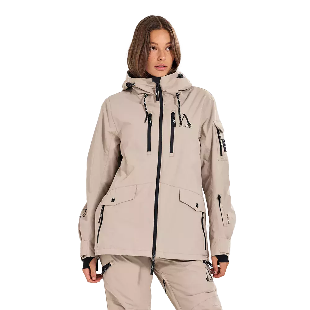 En model iført en lys beige, teknisk outdoorjakke med hætte og sorte kontrastdetaljer. Jakken har to vertikale lynlåse foran, flaplommer med knapper, håndledsmanchetter og justerbare snore i hætten. Matchende beige bukser giver et samlet look. Samlet fremstår outfittet funktionelt og stilrent, velegnet til udendørs aktiviteter med fokus på komfort og praktiske lommer. Detaljerne i sorte lynlåse og snore giver et moderne præg.