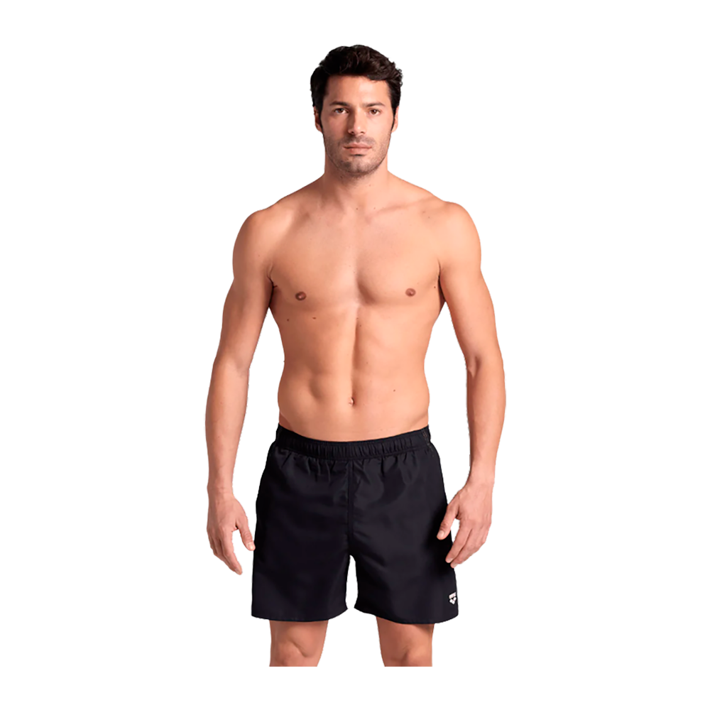 En veltrænet mand står afslappet foran kameraet iført sorte bade- eller træningsshorts. Han er bar overkrop med markeret muskulatur og neutralt ansigtsudtryk, kort mørkt hår og let solbrun hudtone. Kroppen er jævnt oplyst af studiobelysning, og hans holdning er rolig med armene langs siderne. Baggrunden er neutral og fremhæver modellen og tøjet.