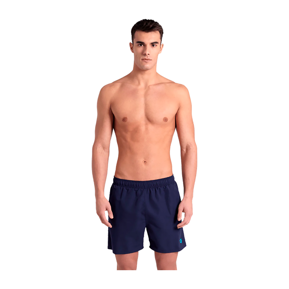 En ung mand står frontalt og afbildet fra hoved til lår på en neutral baggrund. Han er skjorteløs og har en veltrænet, slank overkrop, kort mørkt hår og et neutralt ansigtsudtryk. Han bærer mørkeblå bade- eller træningsshorts med elastik i taljen, hænderne hviler afslappet langs siderne. Billedet fremhæver kroppens symmetri og en enkel, afdæmpet præsentation.