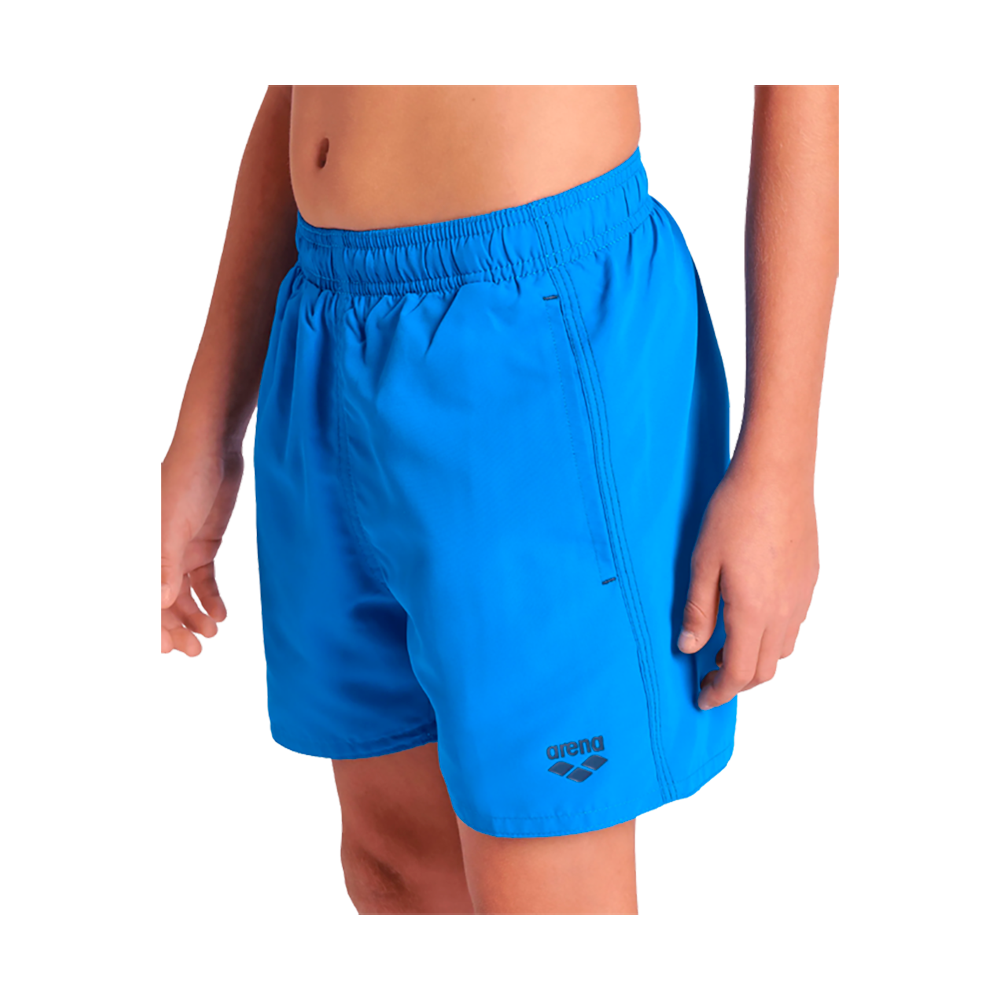 Billedet viser en barn/ung iført korte, klareblå badebukser med elastisk talje. Bukserne har en simpel, sportslig pasform med synlige sidesømme og et lille arena-logo på venstre lår. Overkroppen er bar til navlen, og armene hænger afslappet ned langs siden. Stoffet ser let og hurtigtørrende ud, velegnet til svømning eller strandbrug. Farven er intens og frisk.