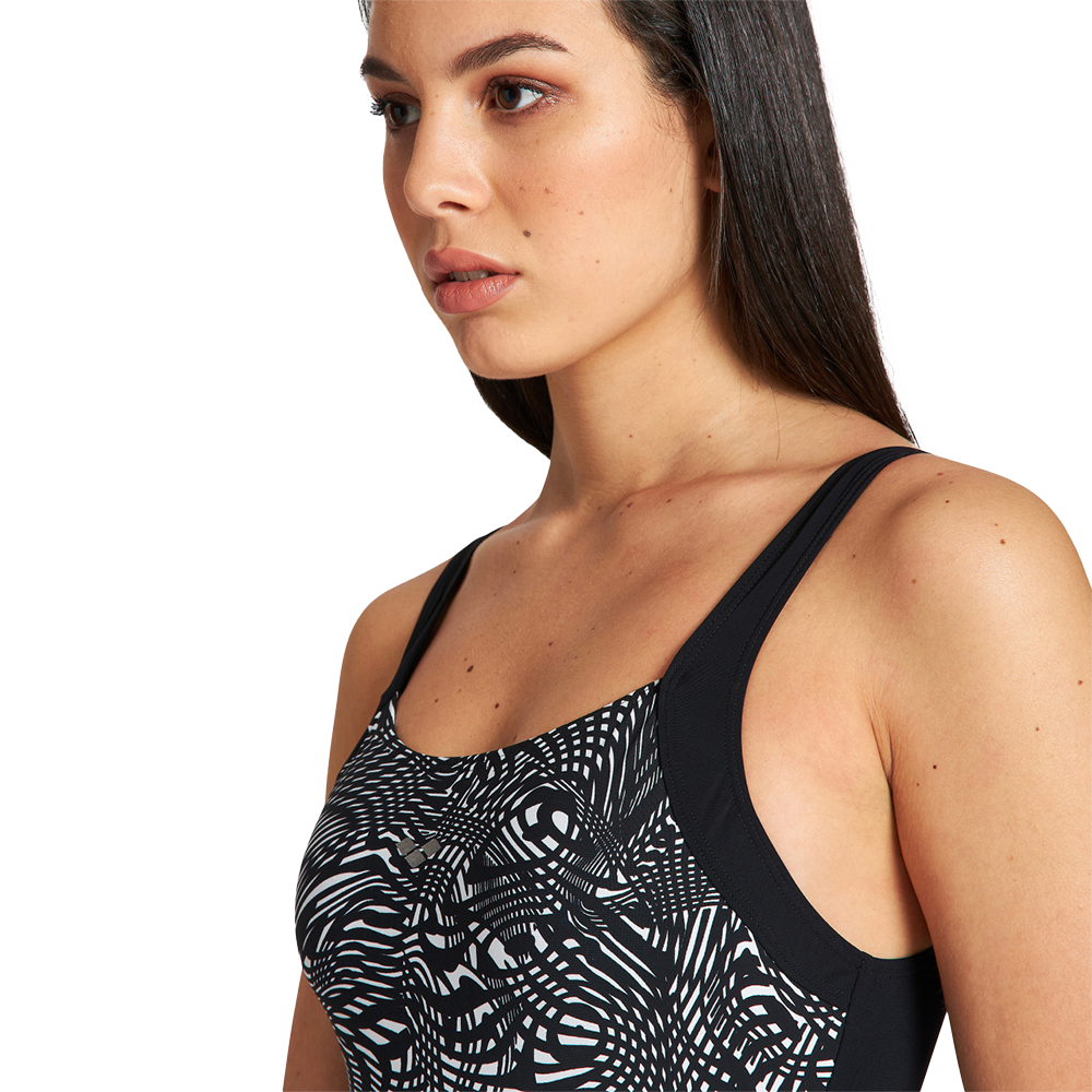 Fotoet viser en ung kvinde i nærbillede fra skuldre og op, iført en ærmeløs badedragt med sort-hvidt, organisk mønster og tynde stropper. Hun har langt, glat mørkt hår, neutral mimik og et fokuseret blik rettet let til venstre. Billedet er taget i studie med hvid baggrund og blødt lys, hvilket fremhæver hudens struktur og mønsterets grafiske kontrast.