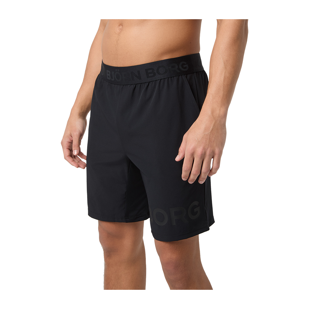 På billedet ses en torso og ben af en person iført sorte træningsshorts fra Björn Borg. Shortsen har en bred elastisk linning med diskret logo og sidelommer. Stoffet virker let og åndbart med en afslappet pasform, der når til over knæene. Personen står i en naturlig, afslappet stilling, og billedet fokuserer på tøjets snit og detaljer.