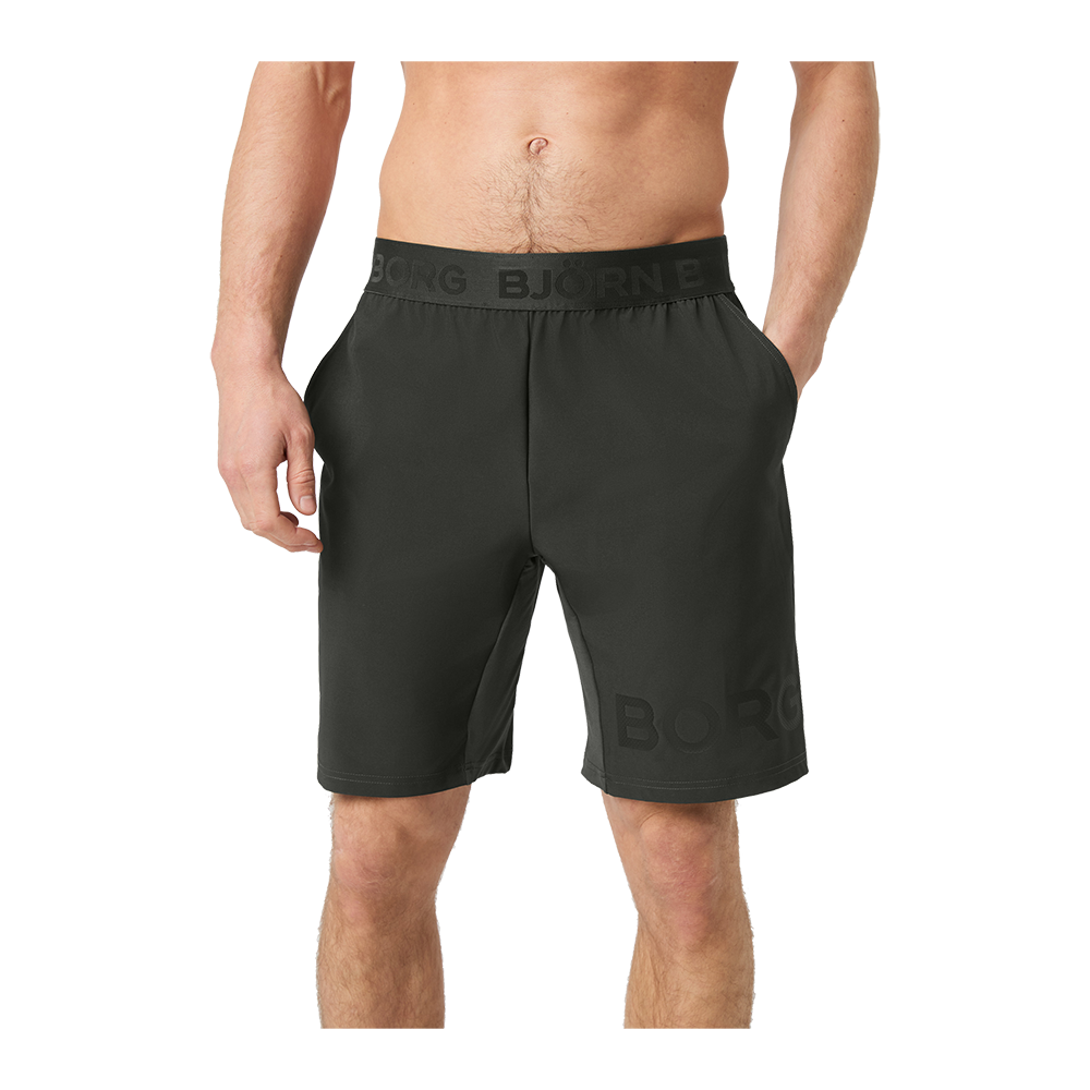 På billedet ses en mandlig model fra taljen og ned iført mørkegrå træningsshorts med elastisk linning og diskret logo. Shortsene har sidelommer og en løs, knælang pasform, som giver bevægelsesfrihed. Stoffet virker let og åndbart, egnet til træning eller afslappet hverdag. Modellen står afslappet med hænderne i lommerne, hvilket fremhæver shortsens komfort og funktionalitet.