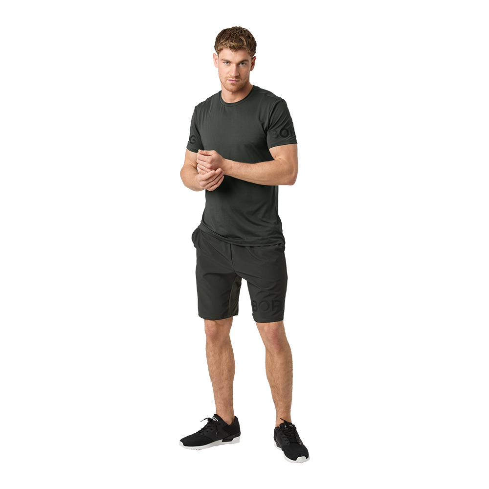 En mand står afslappet i et ensfarvet, mørkt træningssæt bestående af T‑shirt og knælange shorts samt sorte løbesko. Han har en atletisk kropsbygning og kigger fremad med et roligt, selvsikkert udtryk, mens han holder hænderne foran kroppen. Billedet har en neutral baggrund og fokuserer på tøjets pasform og funktionelle udtryk, hvilket giver et simpelt og moderne sportstema.