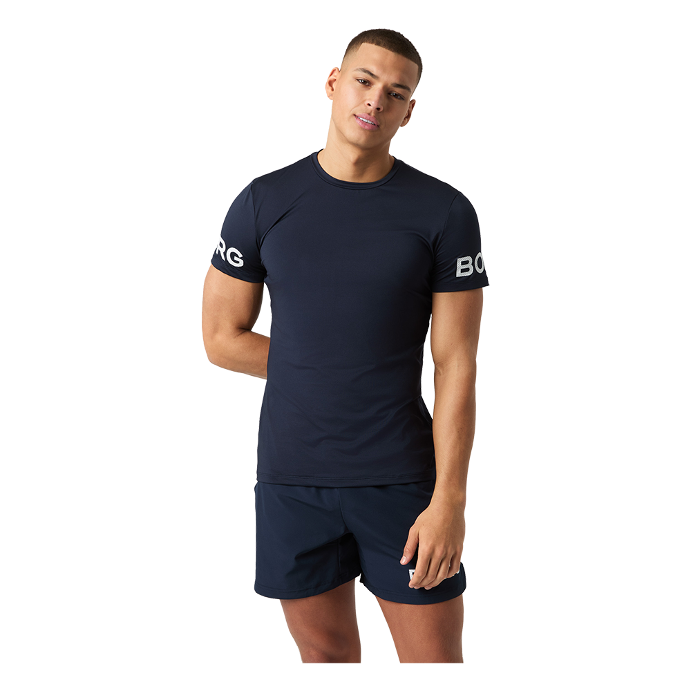 En ung mand står afslappet iført en tætsiddende, mørkeblå trænings-T-shirt og matchende shorts. Han har kort hår, en selvsikker men venlig mine og en let fremadbøjet hovedstilling. Tøjet fremhæver hans atletiske kropsbygning, og poseringen virker naturlig og rolig. Billedet signalerer sport, hverdagstræning og komfortabelt, funktionelt tøj til aktive aktiviteter og bevægelse.