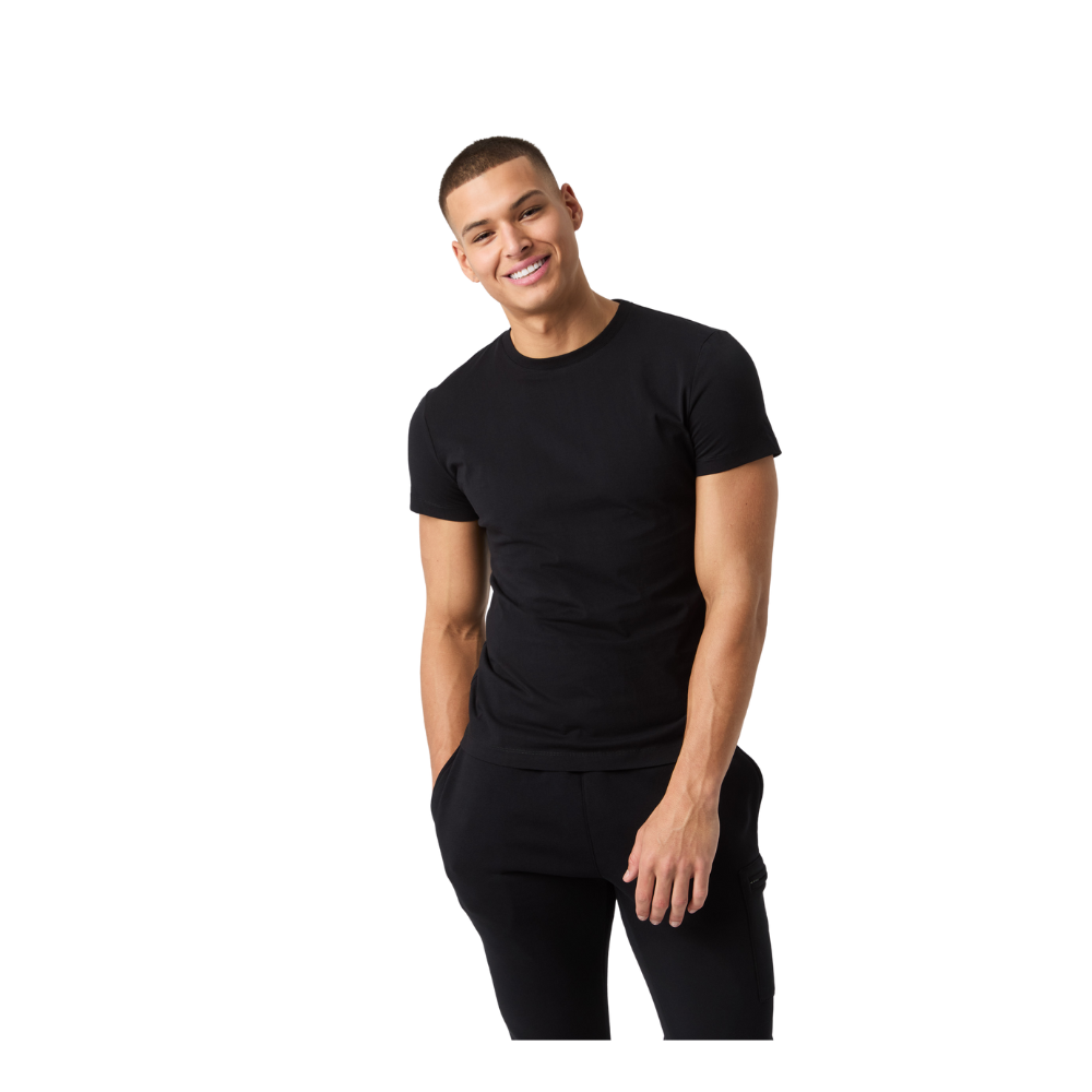 En ung mand står afslappet med hænderne i bukselommerne og et venligt smil. Han bærer en simpel sort t-shirt og mørke bukser, har kort hår og en oprejst, men afslappet holdning. Billedets minimalistiske, neutrale baggrund fremhæver hans stil og naturlige udtryk, hvilket giver indtryk af selvtillid og imødekommenhed og rolig udstråling.