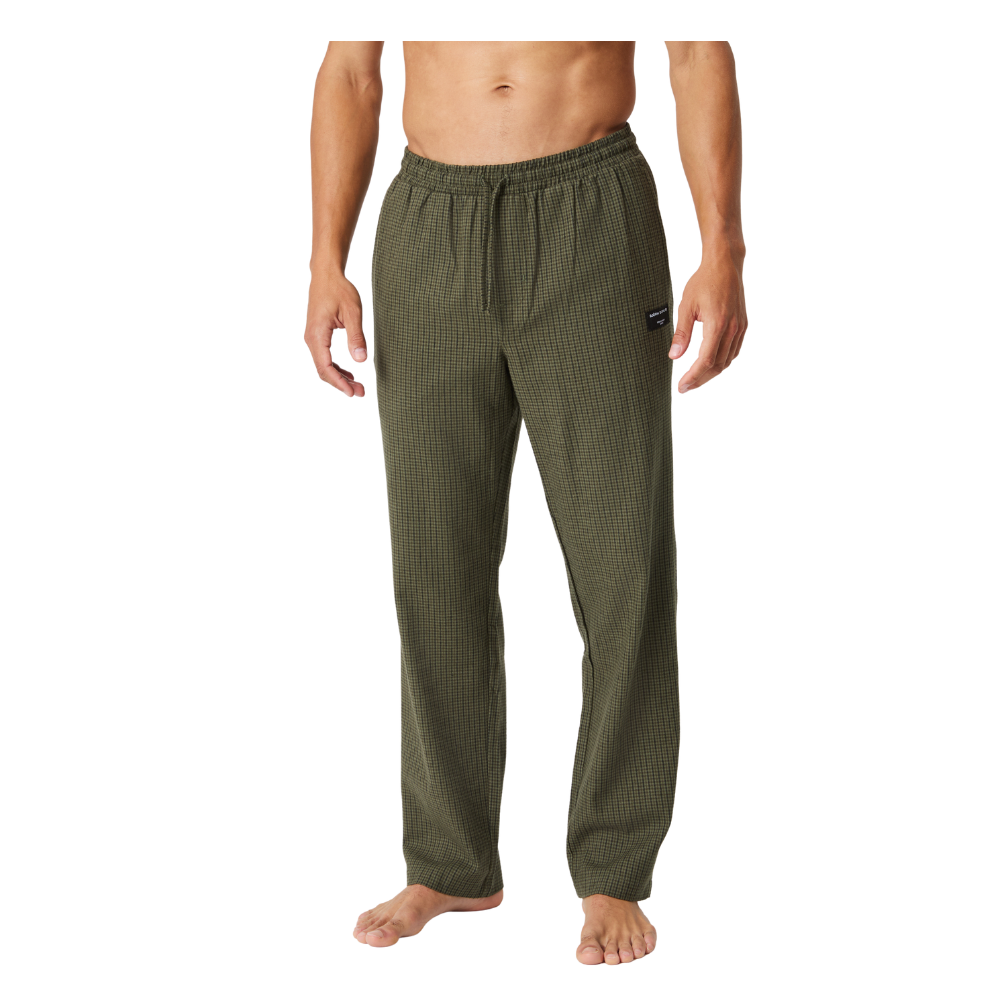 En mand iført afslappede, olivengrønne pyjama- eller loungebukser med en diskret ternet struktur. Bukserne har elastik i taljen og løbegang, sidder løst og komfortabelt langs benene og når næsten ned til fødderne, hvor modellen står eller går barfodet. Stilen signalerer hjemlig hygge og afslapning, egnet til afslappede aftener eller som behageligt tøj til indendørs brug.