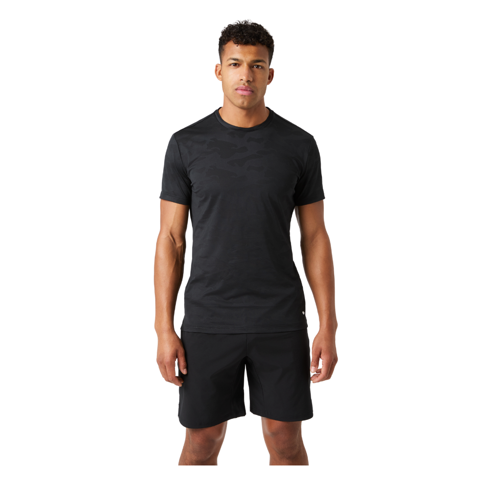 En ung mand står frontalt med en afslappet, rank holdning. Han har kort, krøllet hår og bærer en tætsiddende, sort T-shirt med et diskret camouflagemønster samt sorte træningsshorts. Hans arme hænger løst ned langs siden, og ansigtsudtrykket er neutralt og roligt. Billedet fremhæver en enkel, sporty stil med fokus på funktionelt træningstøj mod en neutral baggrund.