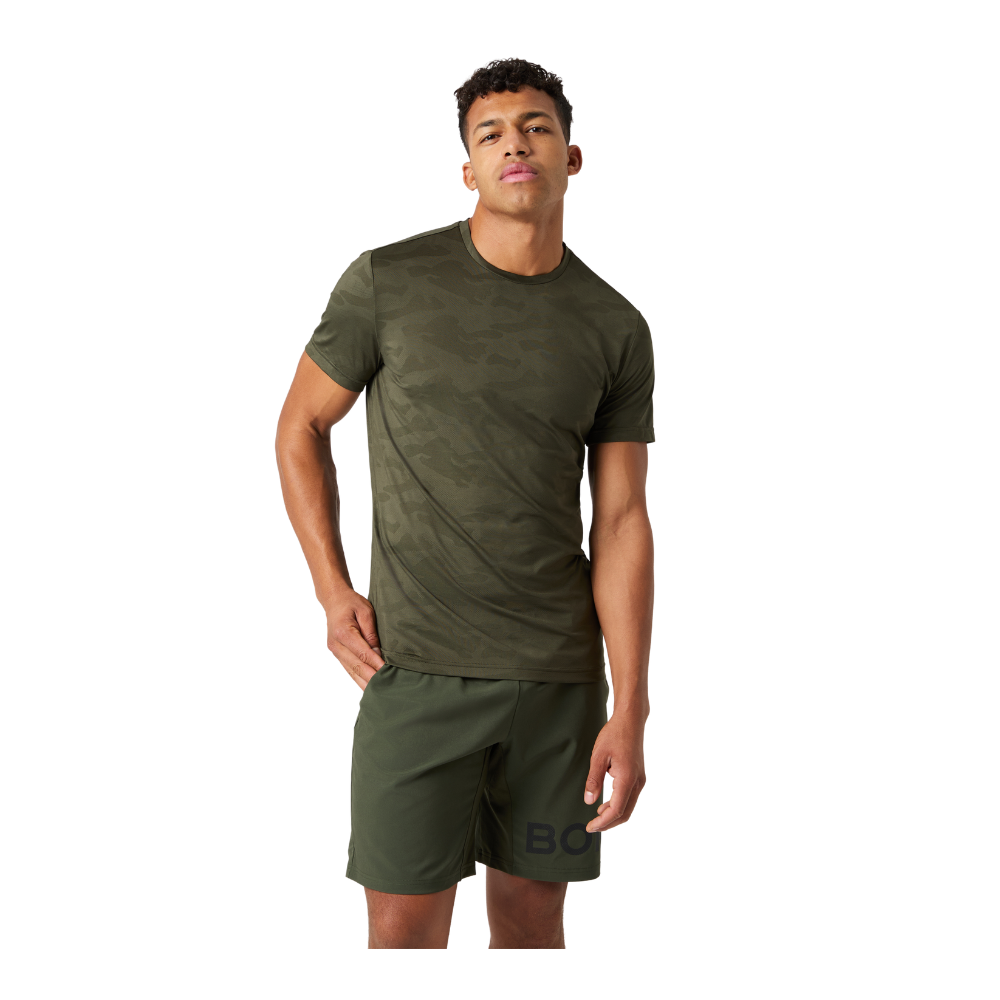 En ung mand står afslappet iført en armygrøn T‑shirt med diskret camouflageprint og matchende træningsshorts. Hans kropsholdning er selvsikker og ubesværet, med den ene hånd i lommen og et let tilbagelænet blik. Outfit'et fremstår enkelt og funktionelt, egnet til både træning og hverdag. Farvetonen giver et råt, moderne udtryk, mens pasformen understreger en sporty, casual stil.