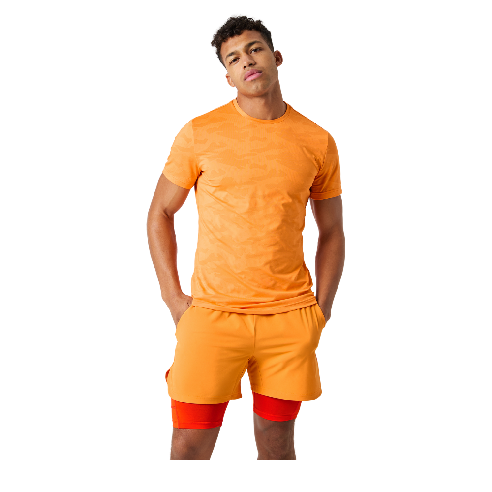 En ung mand står afslappet med hænderne i lommerne iført en knaldorange træningst‑tenue bestående af en tætsiddende t‑shirt og korte shorts med røde indershorts. Han har kort krøllet hår og et roligt, selvsikkert udtryk, mens kroppen er let fremadlænet. Billedet formidler en sporty, moderne og energisk stemning med fokus på stærke farver og en afslappet holdning.