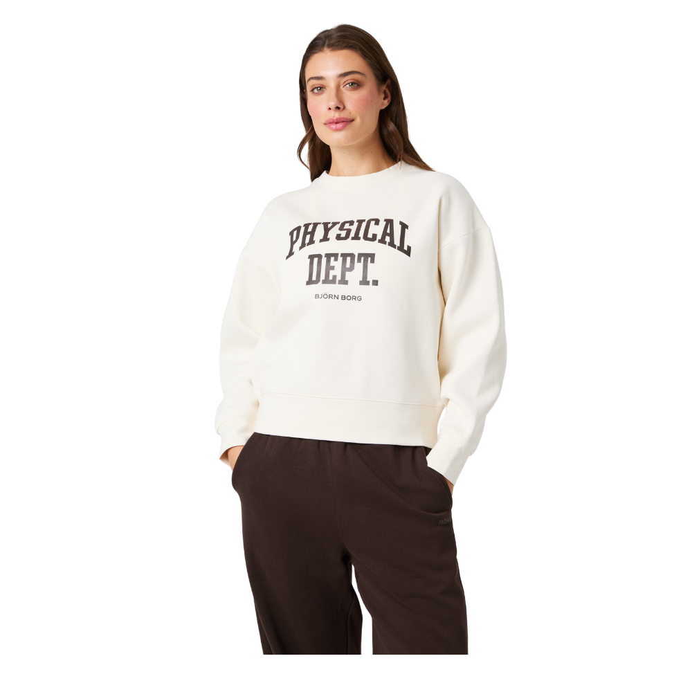 En kvinde står afslappet med hænderne i lommerne iført en cremefarvet sweatshirt med teksten 'PHYSICAL DEPT.' og 'BJÖRN BORG' på brystet. Sweatshirten har en løs, komfortabel pasform og er kombineret med mørkebrune, afslappede bukser. Hendes udtryk er roligt og naturligt, og hele looket signalerer sporty, casual og behagelig hverdagsstil. Billedet virker indbydende og moderne.