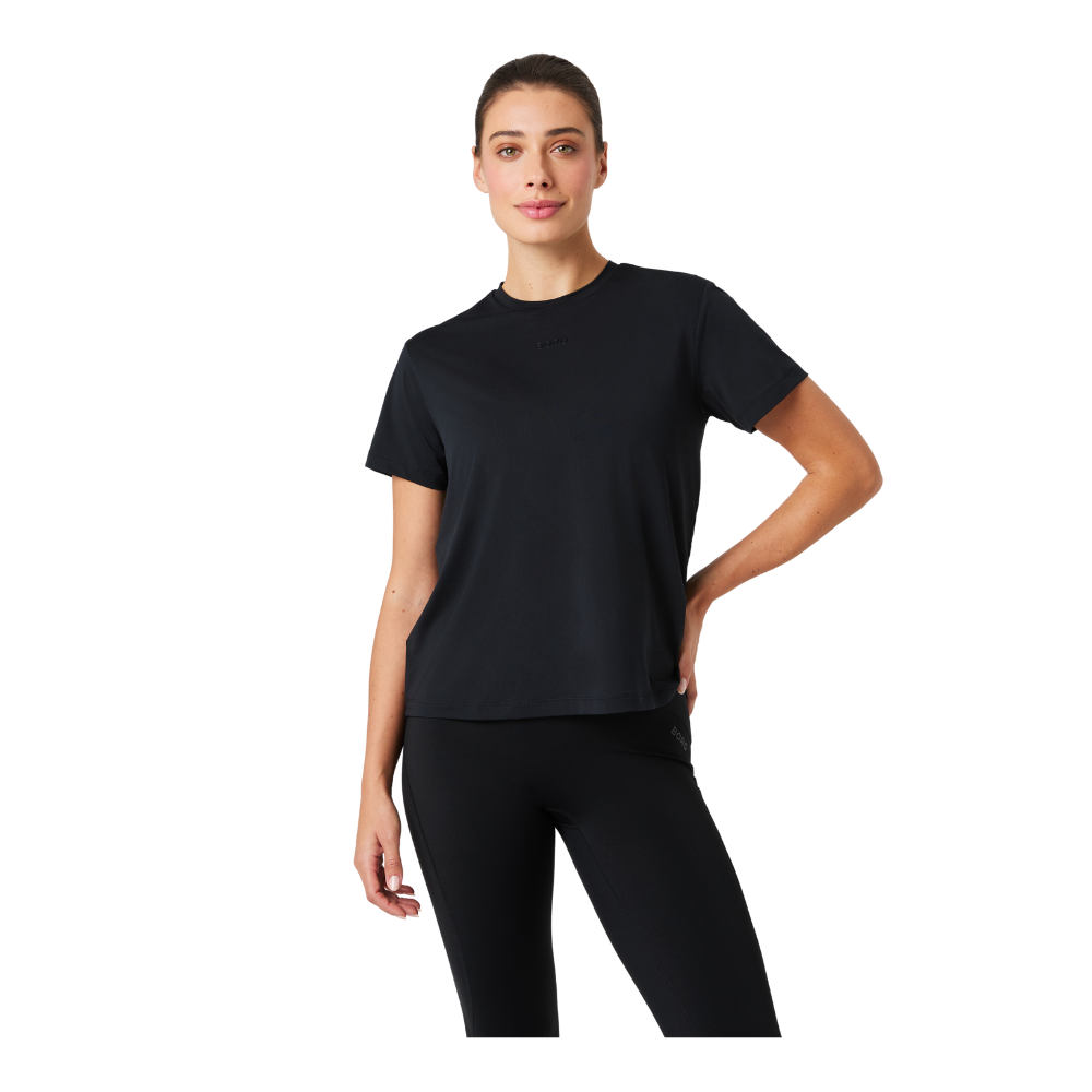 En kvinde står afslappet mod en neutral baggrund iført en enkel, sort t-shirt og tætsiddende sorte træningsleggings. Hun har en rolig, selvsikker holdning med den ene hånd hvilende på hoften og et let smil. Billedet fremhæver et minimalistisk, sporty look med rene linjer, neutral styling og komfortabel pasform, velegnet til både træning og afslappet hverdag.