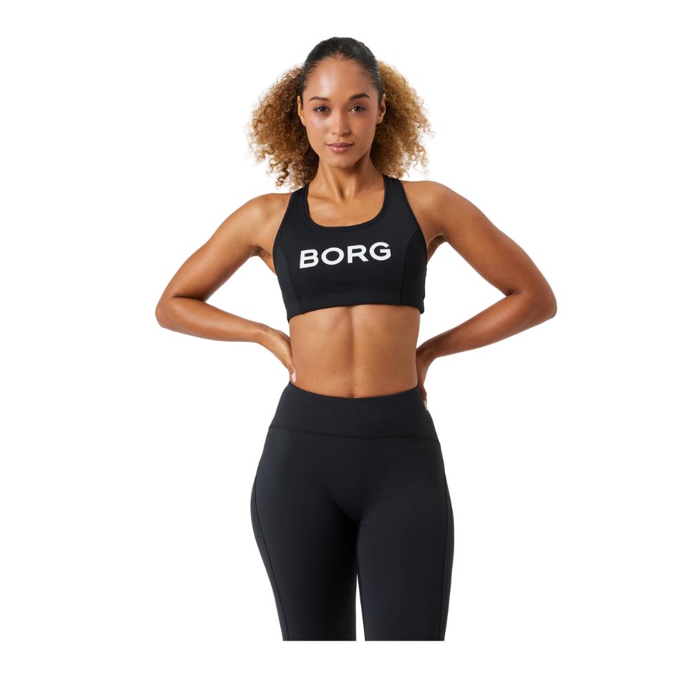 Billedet viser en atletisk kvinde i sort træningstøj med en sports-bh med teksten "BORG" og matchende leggings. Hun står selvsikkert med hænderne i siden, ret holdning og et roligt smil, hvilket giver indtryk af styrke og overskud. Hendes krøllede hår er trukket tilbage, og hendes tonerede mave og arme fremhæver en aktiv livsstil. Stemningen er energisk og inspirerende.