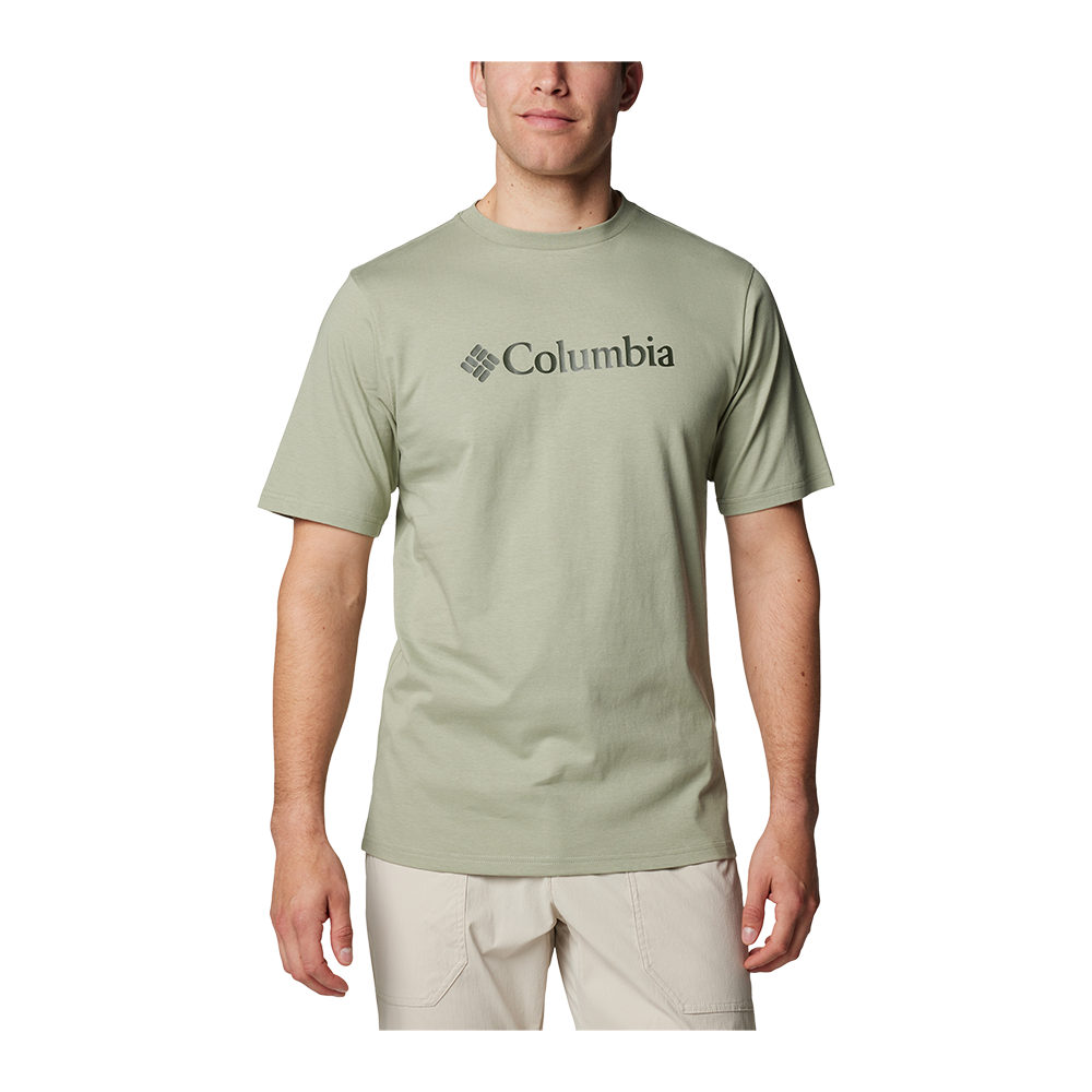 En ung mand bærer en enkel, lysegrøn Columbia T‑shirt med stort logo midt på brystet. T‑shirten har rund hals og korte ærmer i en afslappet pasform og er kombineret med lyse, beige shorts. Billedet har en neutral, hvid baggrund, hvilket fremhæver tøjets farver og snit. Samlet giver udtrykket et enkelt og funktionelt, hverdags- eller friluftsinspireret look.