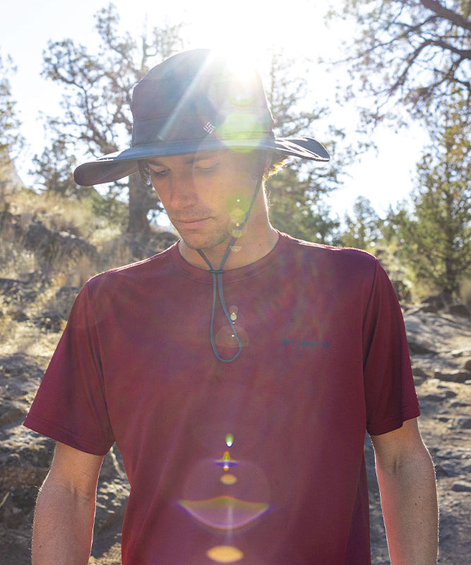 En mand iført en bredskyggelue og en dybrød T‑shirt står udendørs på en tør, stenet skråning omgivet af lave træer. Solen skinner bag ham og skaber stærke lens‑flare-effekter over billedet, mens han kigger ned med et roligt, eftertænksomt udtryk. Stemningen er varm og stille, med skarpt lys, naturlige jordfarver og et indtryk af en vandretur i solskinsvejr.