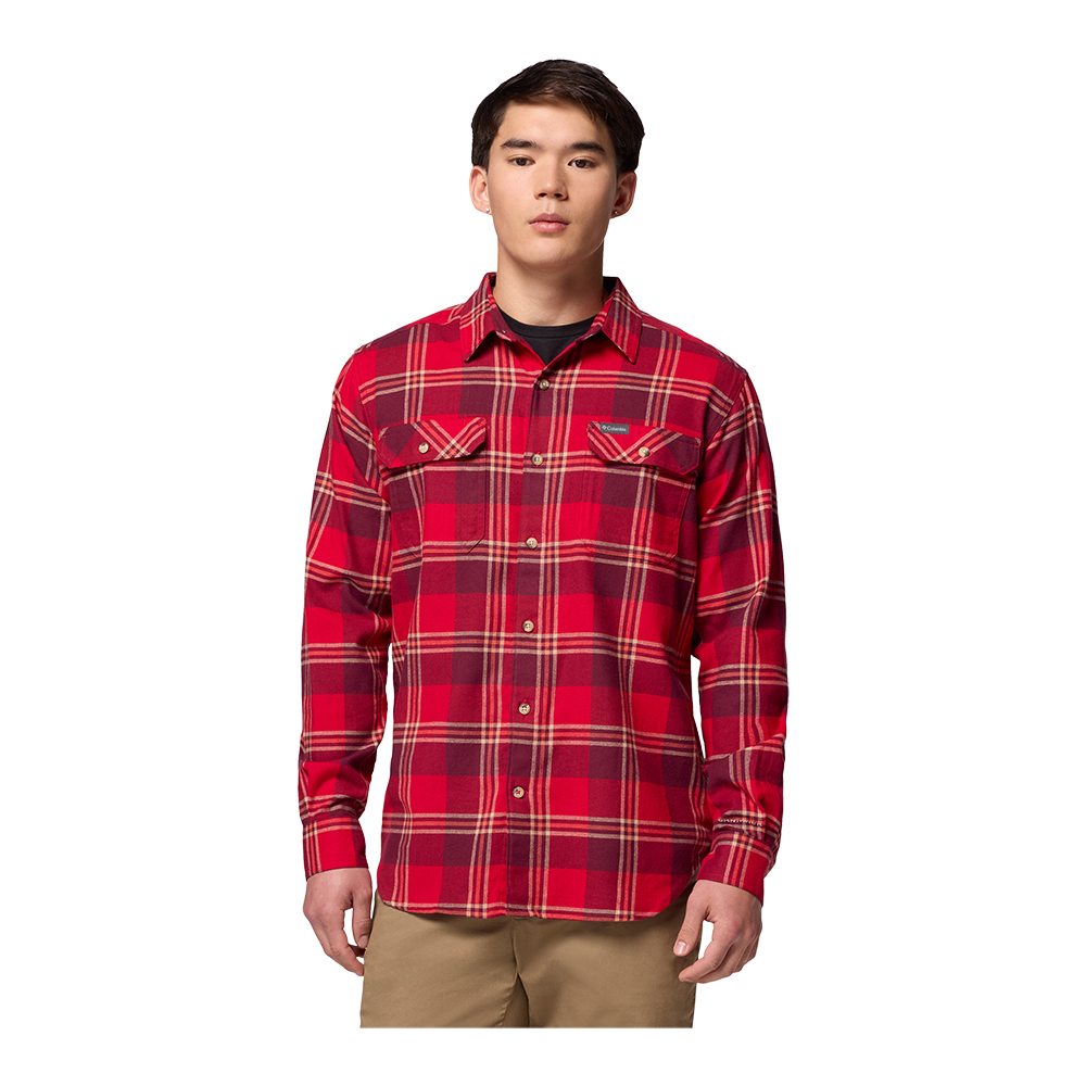 En ung mand står fremadrettet iført en rødternet flannel‑skjorte med knapper og brystlommer, kombineret med beige bukser. Skjorten har varme røde og mørkere tern samt diskrete beige striber. Han har et neutralt, afslappet udtryk og korte, mørke hår. Billedet er enkelt og minimalistisk med hvid baggrund, hvilket fremhæver tøjets farver og snit.