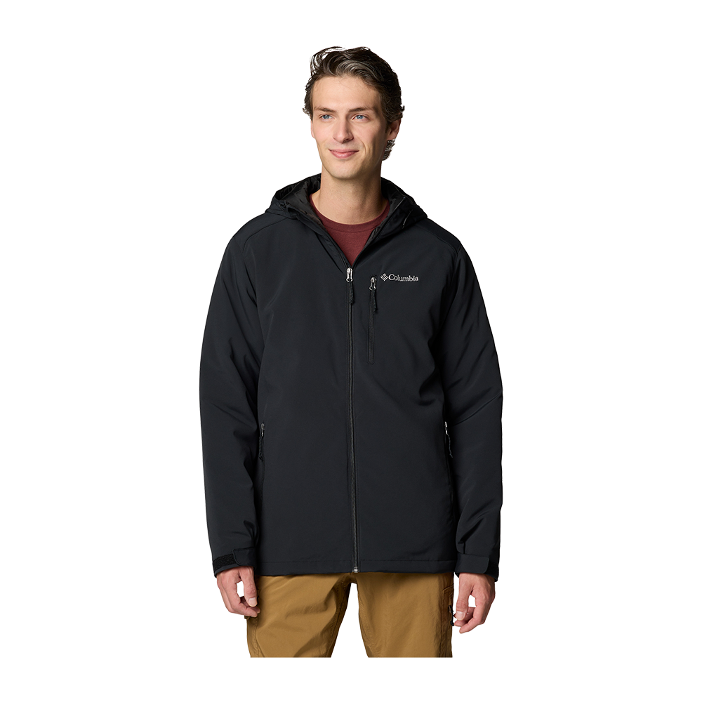En ung voksen mand står afslappet iført en sort Columbia-softshelljakke med hætte, let åbnet lynlås og brystlomme, kombineret med en vinrød t-shirt og beige bukser. Jakken har et diskret logo på brystet og praktiske lynlåslommer. Udtrykket er roligt og naturligt, og billedet fremhæver det funktionelle, hverdagsagtige udendørslook, der både virker praktisk og stilrent.