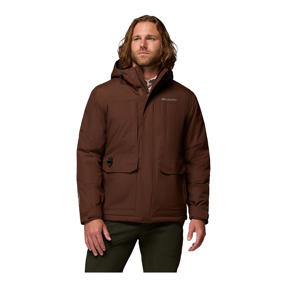 En mand iført en brun, polstret vinterjakke med hætte står i afslappet positur. Jakken har to store forlommer med klap, justerbare manchetter og en skjult lynlås med trykknapslukning. Materialet virker robust og vandafvisende, egnet til kolde, våde forhold. Udtrykket er neutralt og praktisk, med et design der kombinerer funktionalitet og tidløs, uformel stil.