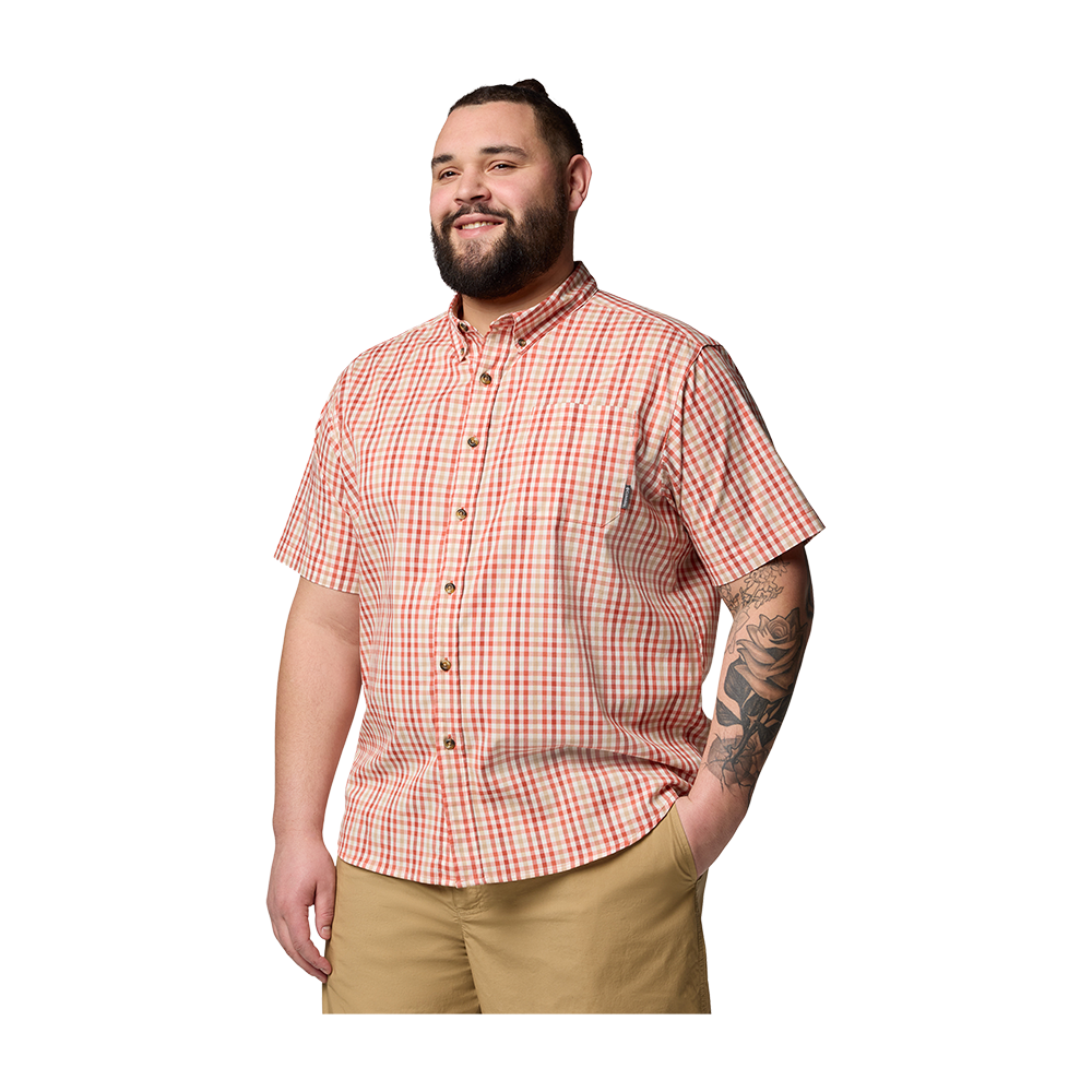 En smilende mand i en kortærmet ternet skjorte og khakifarvede shorts står afslappet med den ene hånd i lommen. Han har kort hår, fyldigt skæg og en iøjnefaldende tattovering af en rose og geometriske elementer på underarmen. Kompositionen giver et venligt og afslappet indtryk, velegnet til fritids- eller sommerstil og casual modepræsentationer.