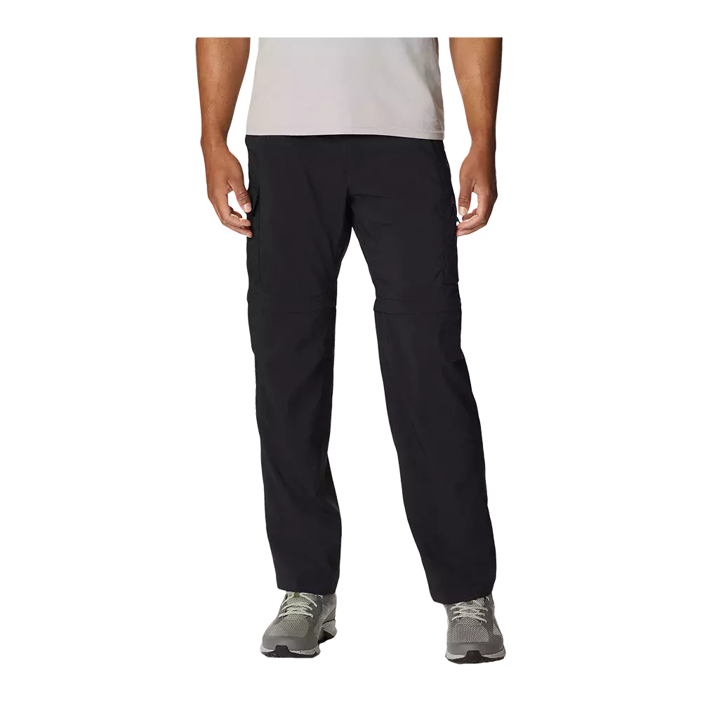 Mandens bukser i billedet er sorte, løstsiddende fritids- og vandrebukser med en lige pasform og praktiske lommer. De ser ud til at være lavet i et let, hurtigtørrende materiale og har en synlig lynlås midt på låret, der antyder aftagelige ben. Bukserne er stylet med grå løbesko og en lys T-shirt, hvilket giver et funktionelt og afslappet outdoor-look.