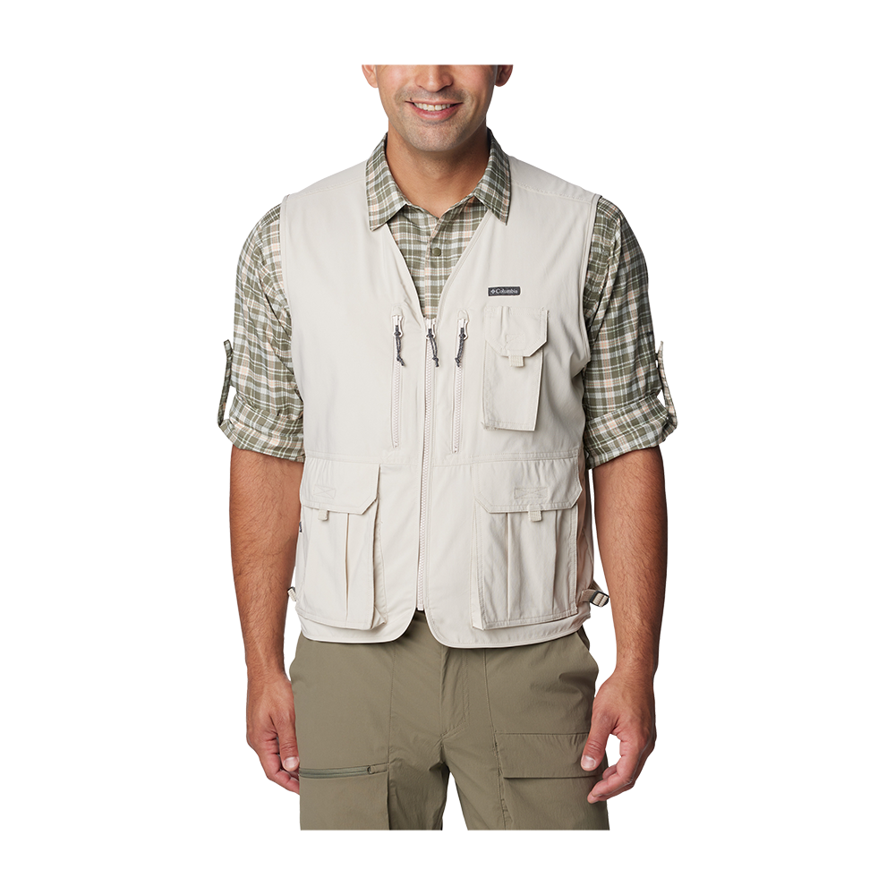 Et nærbillede viser en mand iført praktisk friluftstøj: en lys beige vest med flere lynlåse- og klaplommer over en grønternet, opkærmet skjorte og olivengrønne cargobukser. Vesten har funktionelle detaljer til opbevaring, mens skjorten giver et afslappet, udendørs præg. Personen står let smilende med hænderne afslappet ned langs siderne, hvilket signalerer en rolig og forberedt attitude til udendørsaktiviteter.