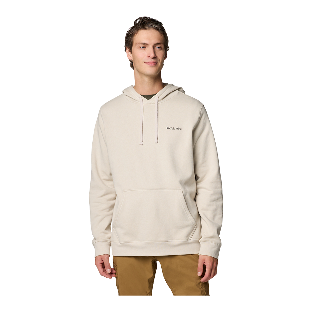En ung mand står afslappet foran en neutral baggrund iført en lys beige hættetrøje med lille Columbia-logo på brystet og brune bukser. Trøjen har snore i hætten og en kængurulomme, og stoffet fremstår blødt og varmt. Hans holdning er rolig med et let smil og hænderne ned langs siden. Billedet formidler en enkel, afslappet og funktionel hverdagsstil.