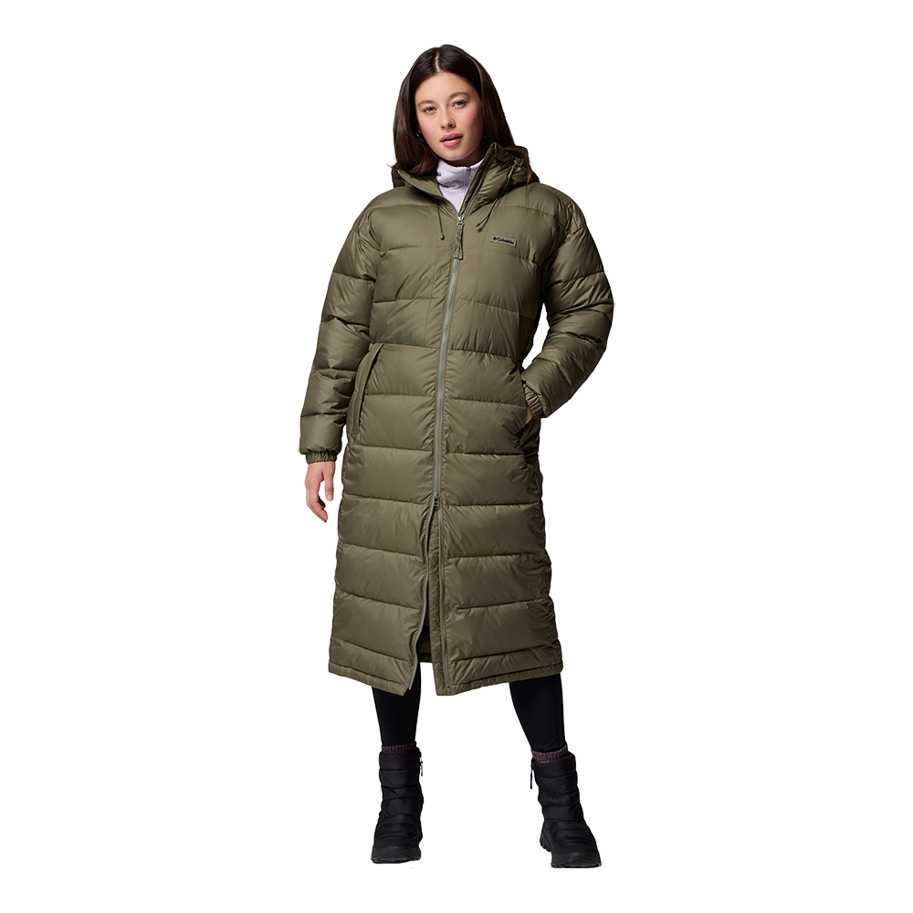 En kvinde står i en lang, olivengrøn dunfrakke med hætte og synlige vandrette quilt-sømme. Frakken når næsten til anklene, har en gennemgående lynlås, indbyggede sidelommer og en afslappet, varm silhuet. Under bærer hun sorte leggings og vinterstøvler, hvilket skaber et praktisk og komfortabelt vinterlook. Modellen har en rolig, neutral holdning, som fremhæver jakken som billedets hovedfokus.