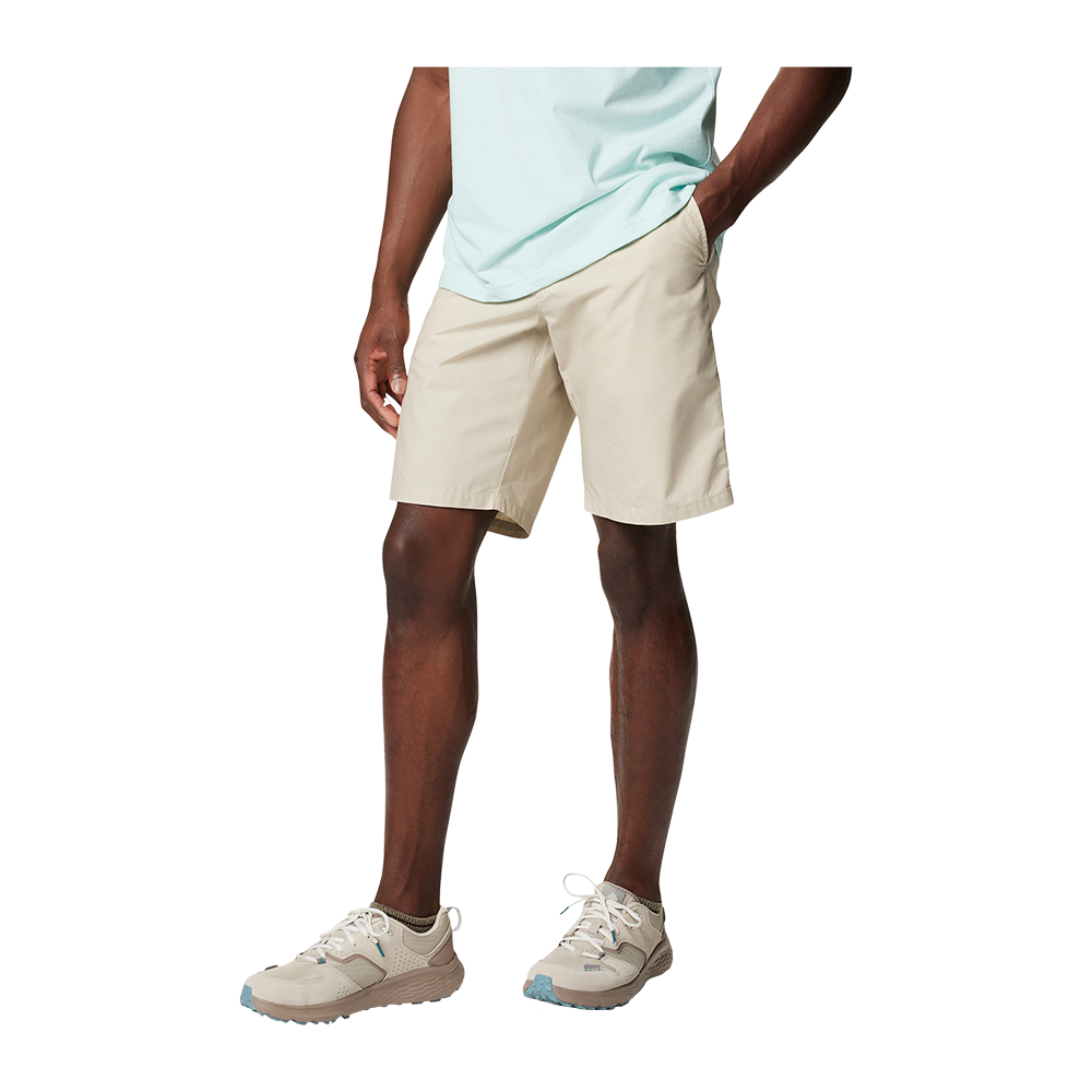 Et stilrent billede af underkroppen på en person iført lyse beige shorts og en mintgrøn t-shirt. Hænderne hviler afslappet i lommerne, mens mørke, veltrænede ben står i en naturlig positur. Fodtøjet er lyse sneakers med diskrete blå accenter, som matcher det lette, sommerlige udtryk. Baggrunden er neutral, hvilket fremhæver tøjets rene, casual stil.