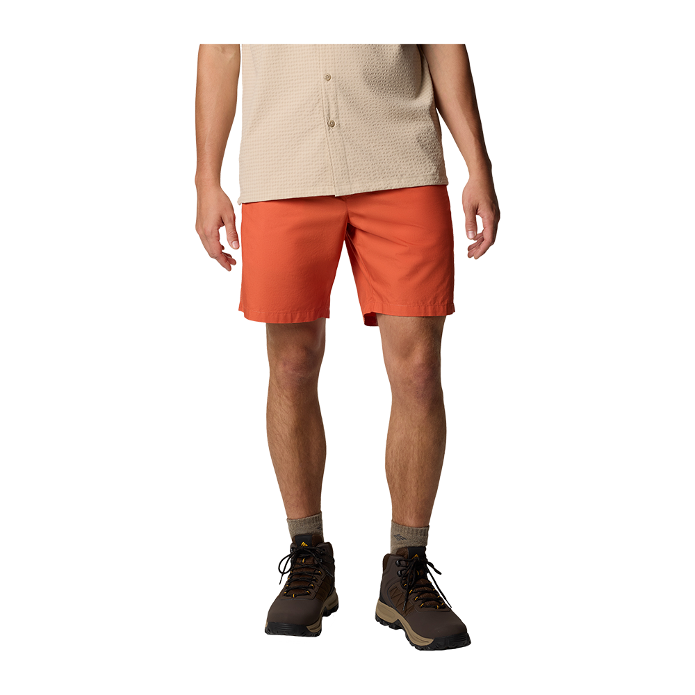 Billedet viser en person klædt i en afslappet, udendørs stil med orange shorts og en beige kortærmet skjorte i våffelstruktur. Benene er bare ned til knæene, iført korte beige sokker og robuste brune vandrestøvler med snørebånd. Kompositionen er enkel og fokuserer på tøj og fodtøj, med neutrale farver og et funktionelt udseende velegnet til sommerlige vandreture eller fritidsaktiviteter.