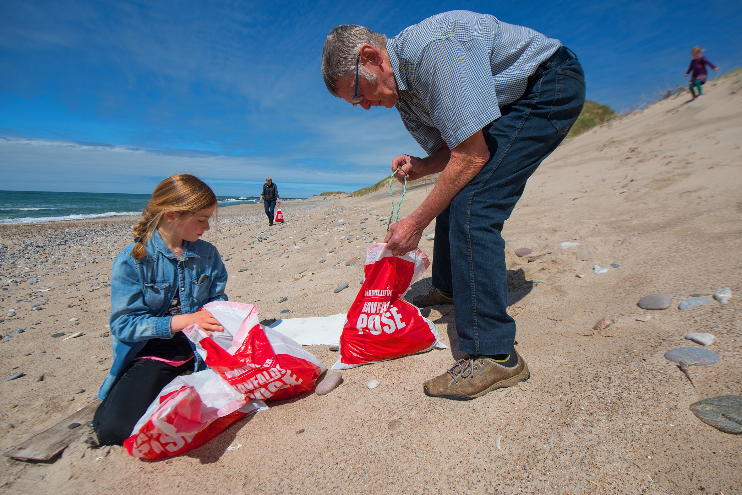 På en vindblæst sandstrand ved Vesterhavet samler en ældre mand og en ung pige affald i røde poser. De arbejder koncentreret med at samle plastik og skrald mellem sten og tang, mens blå himmel og rolige bølger danner baggrund. Et par andre personer går langs kysten i det fjerne. Situationen udstråler omsorg for naturen og fælles ansvar på tværs af generationer.