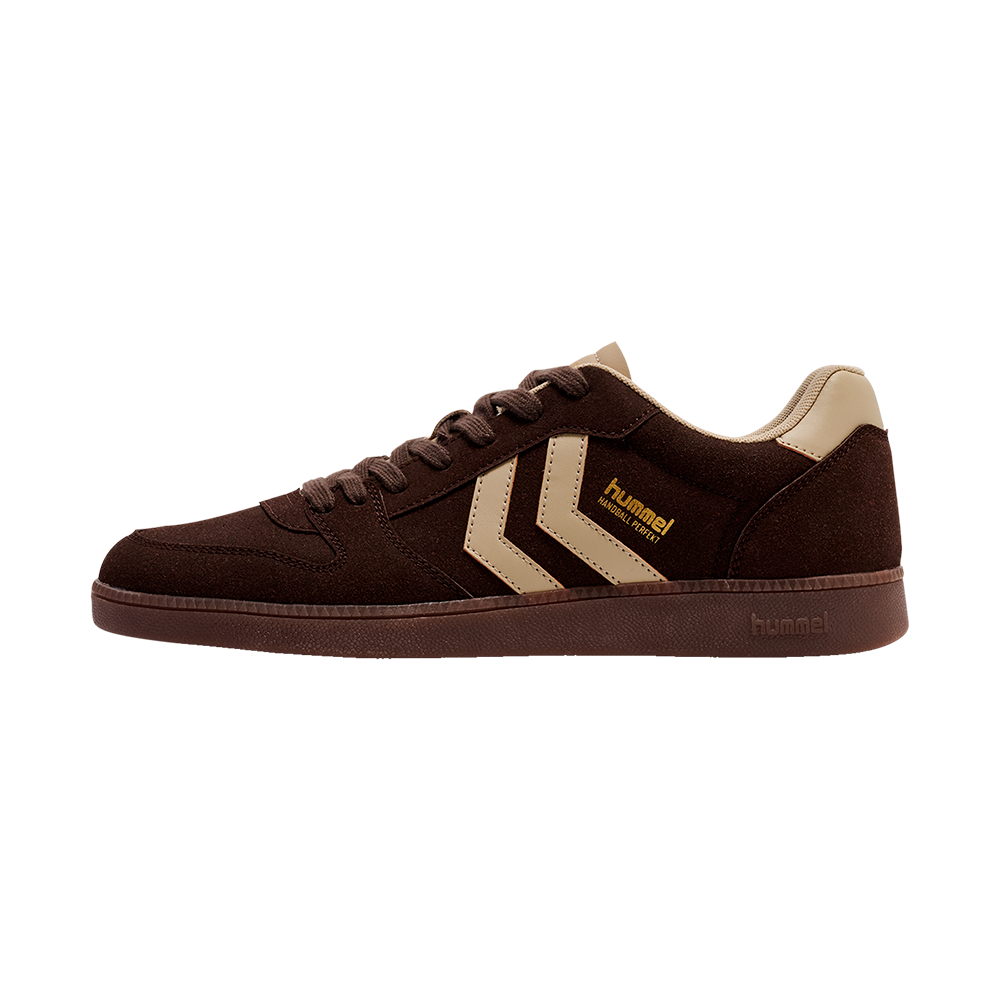 Et stilrent, lavprofil sneakers i mørkebrunt ruskind med beige detaljer. Skoen har flade snørebånd, en brun gummisål og to beige chevron-striber på siden, hvor det lille "hummel" logo fremgår i gul tekst. Hælparti og indlæg er polstrede og lysebeige, hvilket skaber kontrast. Designet virker enkelt og robust, egnet til hverdag og afslappede outfits.