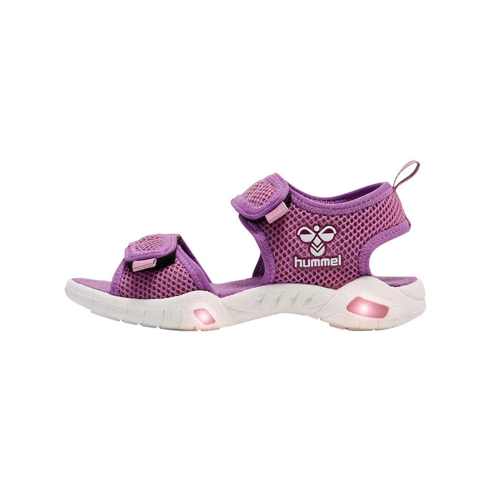 Sandalen er en lilla børnesandal fra Hummel med åndbart mesh og velcrolukninger for justerbar pasform. Den har to brede remme over foden og en støtte ved hælen, samt det genkendelige Hummel-logo på siden. Den hvide, bløde sål har indbyggede LED-lys, der giver et sjovt, blinkende element. Designet kombinerer komfort, ventilation og legesyg stil til børn.