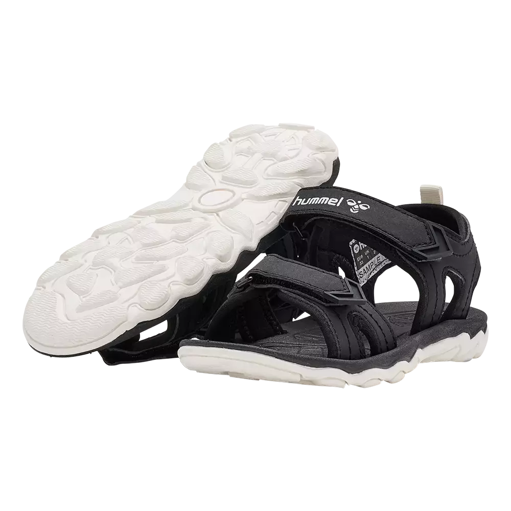 På billedet ses et par sorte sportssandaler fra Hummel med kraftig, hvid gummisål og markante mønstre til greb. Sandalerne har justerbare velcroremme over vristen og ved anklen, åben tå og sidestøtte for god ventilation. Indersålen virker polstret for komfort, mens den robuste ydersål giver stabilitet på ujævnt terræn. Designet kombinerer funktionalitet og et enkelt, moderne udtryk til sommerbrug.