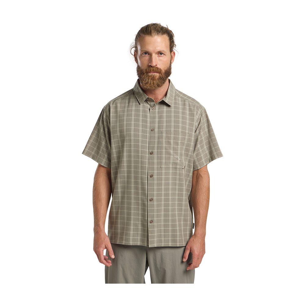 En voksen mand står foran kameraet med et roligt, neutralt ansigtsudtryk. Han har langt hår trukket tilbage i en knold og et fyldigt, rødlige skæg. Han bærer en kortærmet, ternet skjorte i afdæmpede jordfarver og lyse bukser, hvilket giver et afslappet, udendørs-aktigt udtryk. Armene hænger afslappet ned langs siden, og den rene, lyse baggrund fremhæver hans afslappede, naturlige stil.