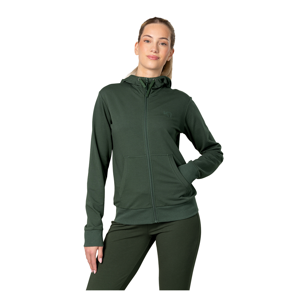 Billedet viser en kvinde iført en mørkegrøn, lynlåstrøje med hætte og matchende leggings. Hun står afslappet med den ene hånd i lommen og et let smil, mens håret er trukket tilbage. Tøjet har rene linjer, ribkanter og en praktisk kængurulomme, hvilket giver et sporty og komfortabelt udtryk egnet til træning eller hverdag.